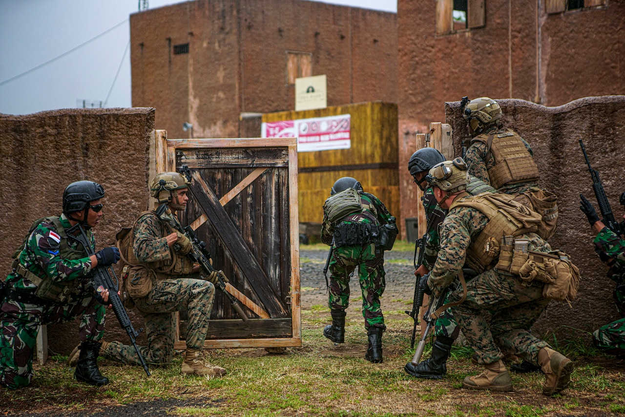 U.S. and Indonesian marines crouch and kneel by a gate outside a building.