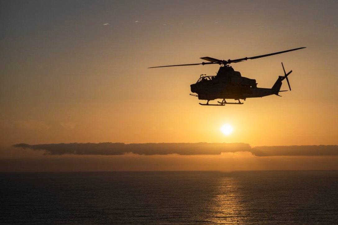 A helicopter flies over water as the sun shines in the background.