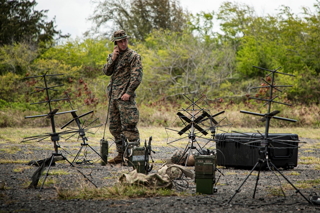 A Marine stands in a field surrounded holding a phone receive and surrounded by numerous antennae.