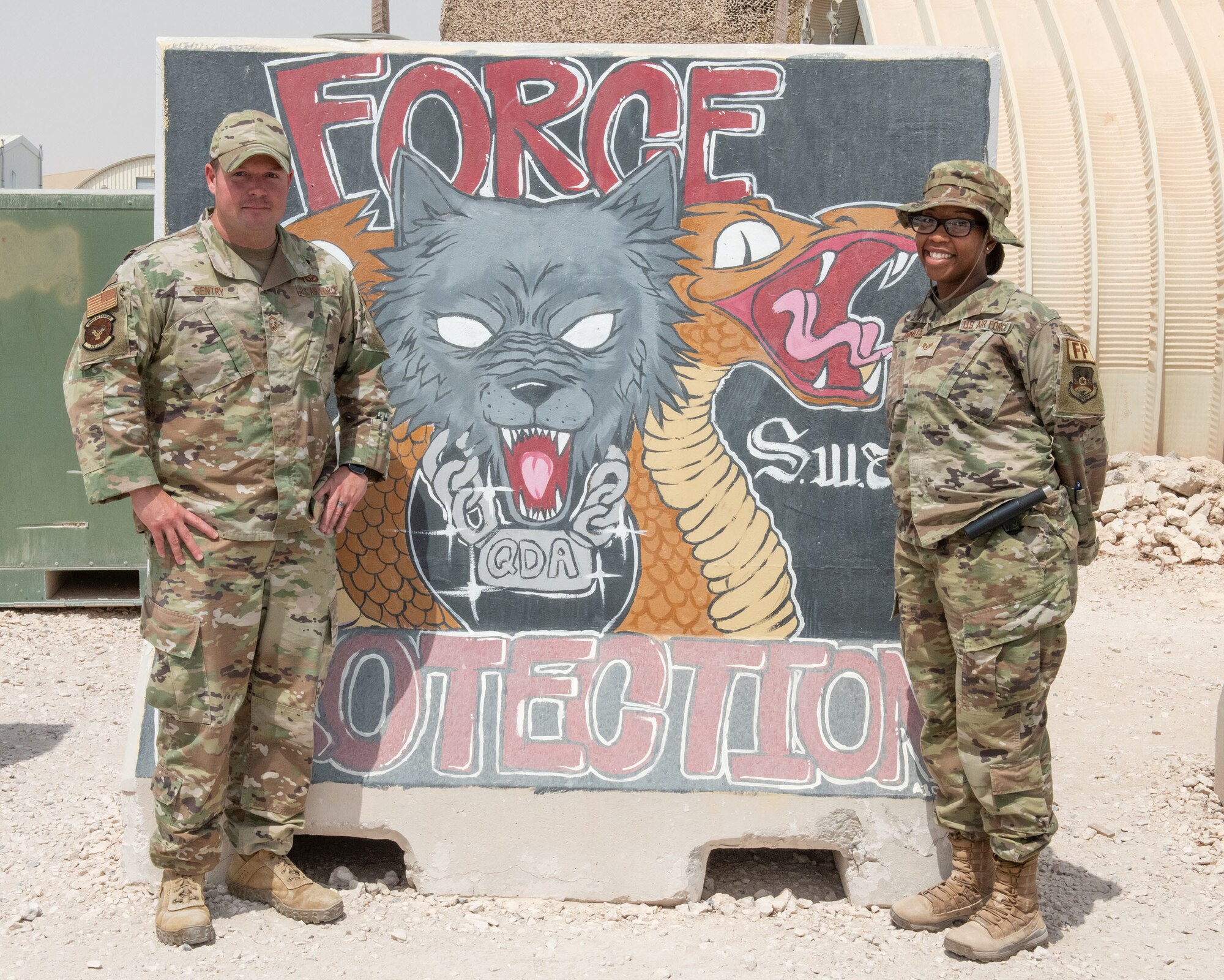 U.S. Air Force Senior Master Sgt. Garret Gentry and Staff Sgt. Briana Bonets, Force Protection airmen with the 379th Expeditionary Civil Engineer Squadron, pose in front of a concrete wall at Al Udeid Air Base, Qatar, July 17, 2022. Force Protection is responsible for the escort and supervision of other-country nationals employed on AUAB. (U.S. Air National Guard photo by Airman 1st Class Constantine Bambakidis)
