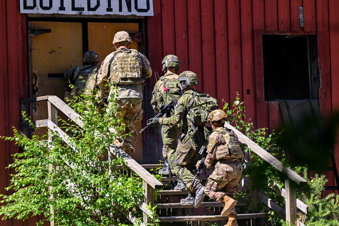 Soldiers carrying weapons walk up steps to a red building.