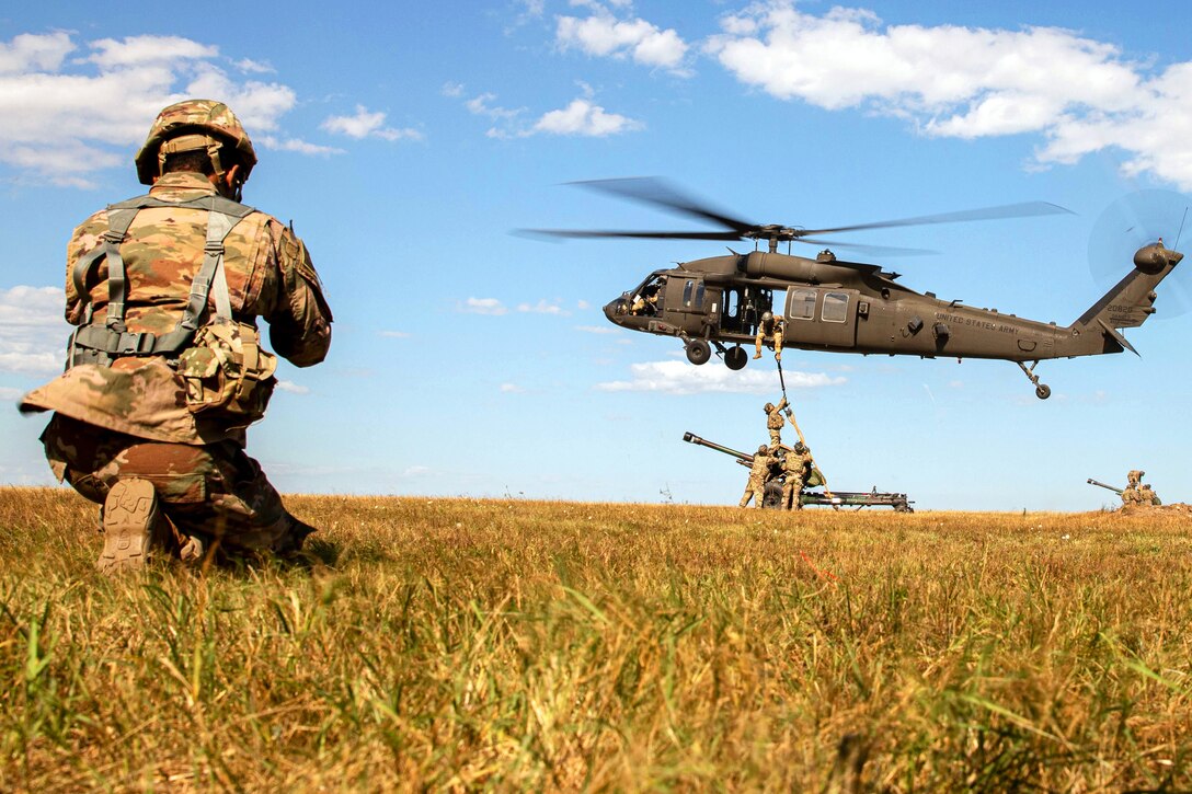 A soldier takes a knee in a field while watching other soldiers in the distance attach a weapon to a hovering helicopter by sling.