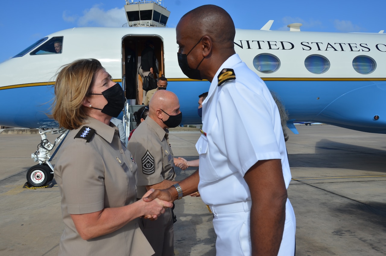 A man and a woman, each in military uniforms, greet each other on the tarmac. In the background is an airplane with the words “United States” on the fuselage.