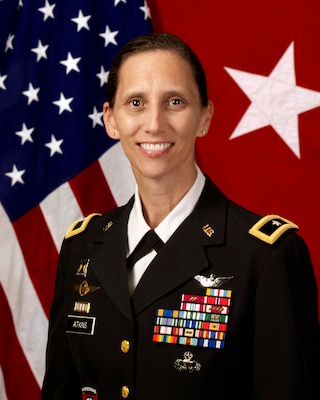 A woman in Army Dress Uniform in front of the US Flag and a One Star General Flag.