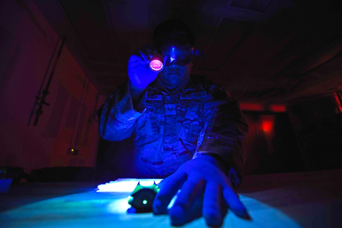 An airman illuminated by blue light uses a flashlight to look at a piece of equipment.