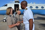 A man and a woman, each in military uniforms, greet each other on the tarmac. In the background is an airplane with the words “United States” on the fuselage.