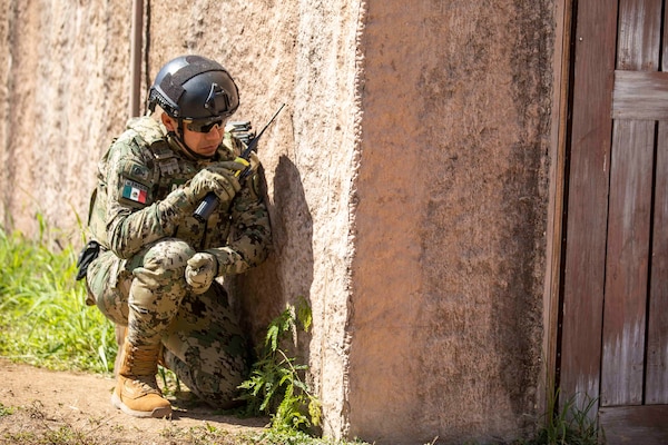 A man in a Mexican military uniform is crouched near a wall, talking on a hand-held radio, during a training exercise.