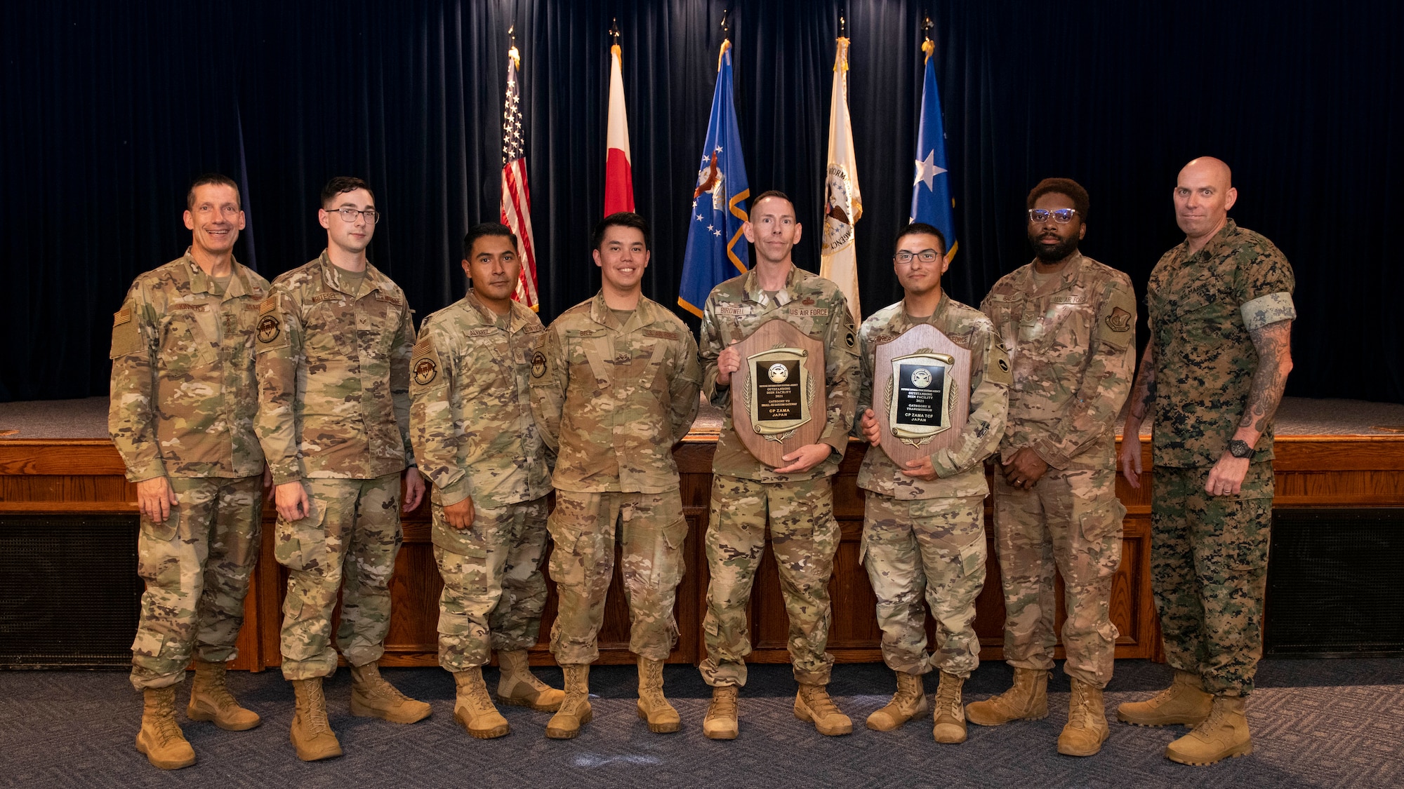 Eight military uniformed men stand next to each other while two of them hold up large wooden award plaques.