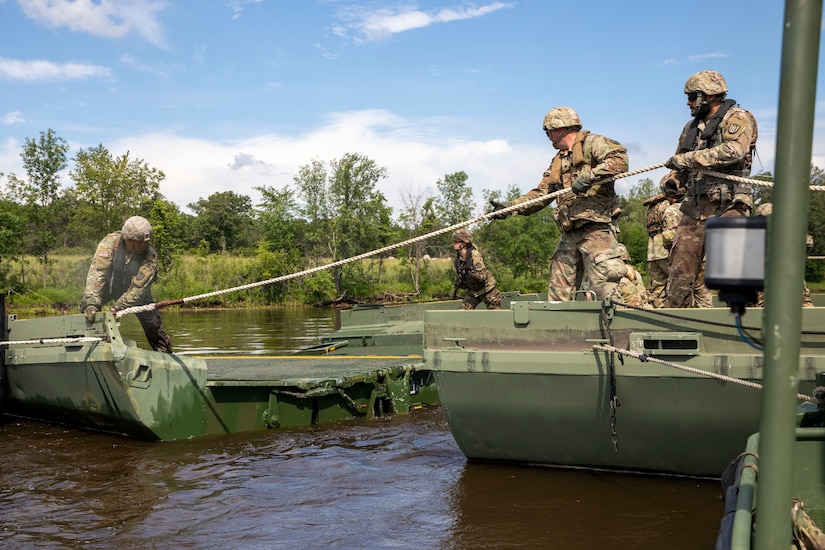 Two soldiers stand on one section of floating bridge pull a rope held by a soldier on another section.