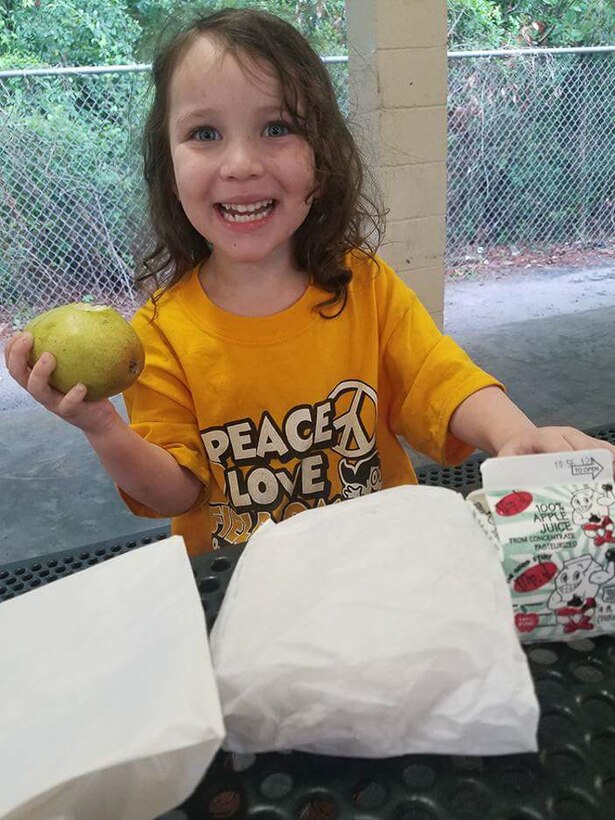 A child holds up a piece of fruit and smiles at a table.
