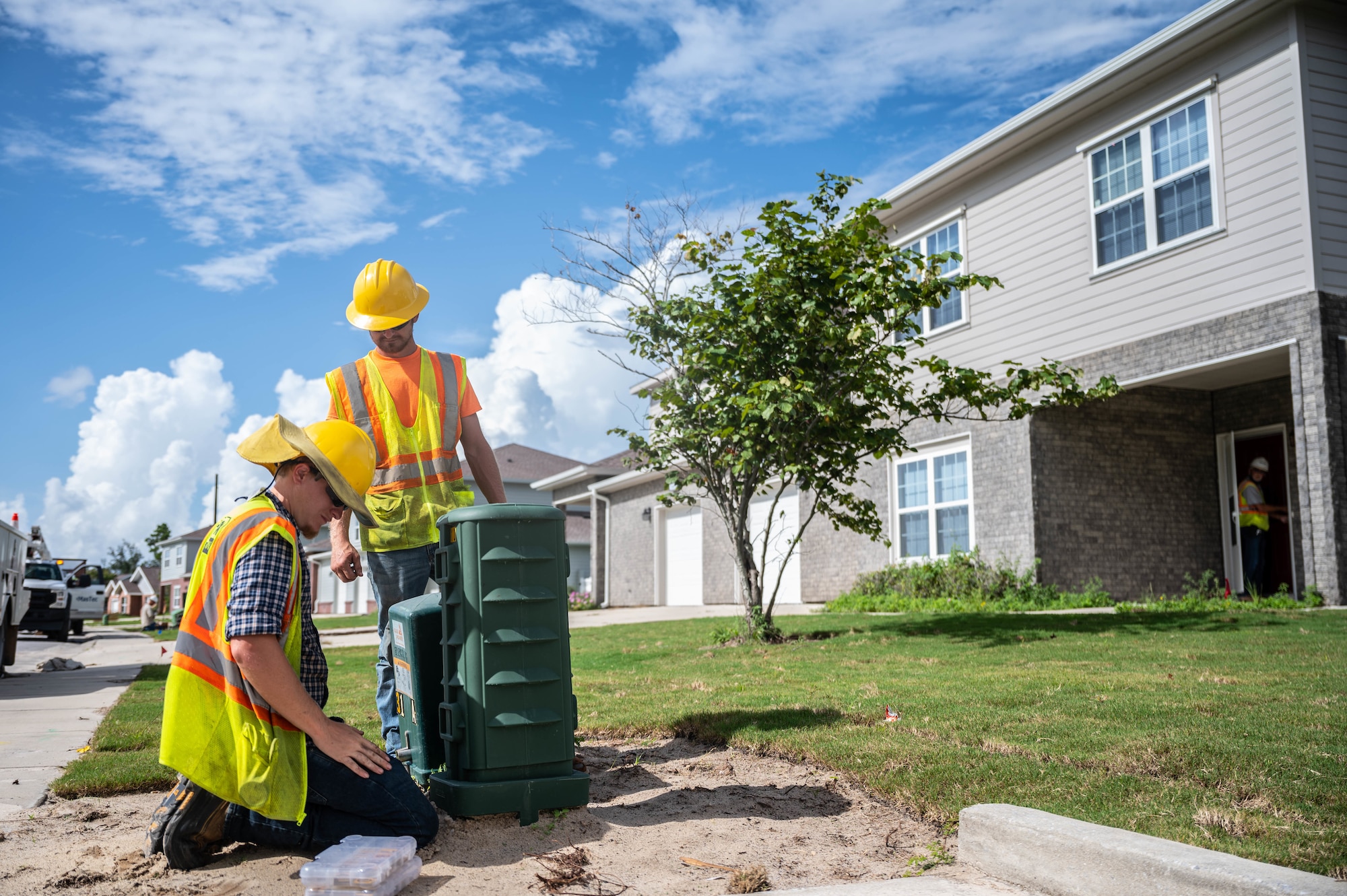 Two men in hard hats install an electrical box outside of a home.