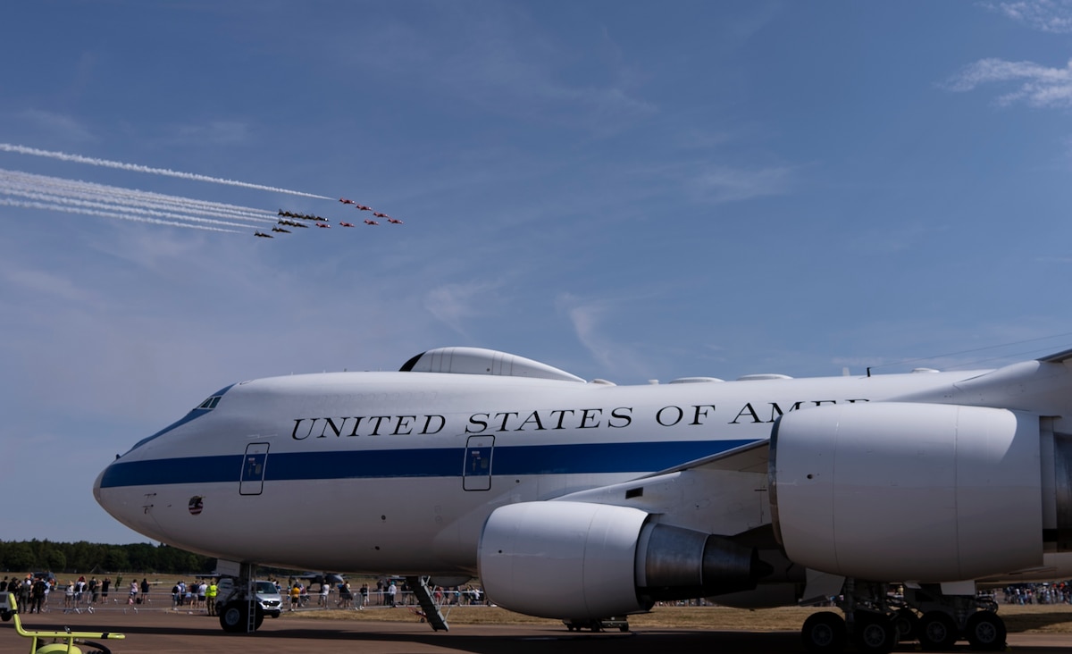 An E-4B sits on display on a tarmac as other aircraft fly over in formation in the background.