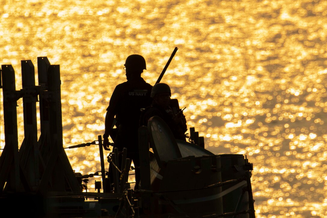 Two sailors shown in silhouette stand aboard a ship.