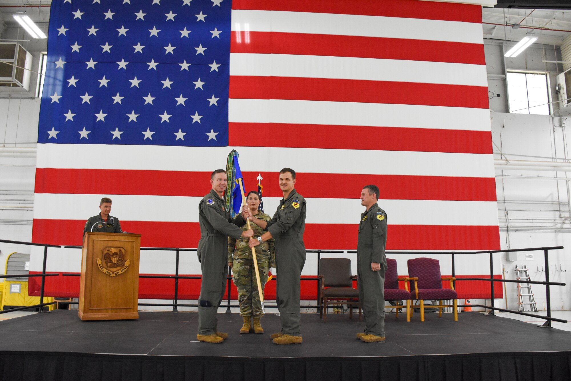 A man hands another man a guidon to symbolically transfer command of the squadron. The two are standing in front of a large American flag.