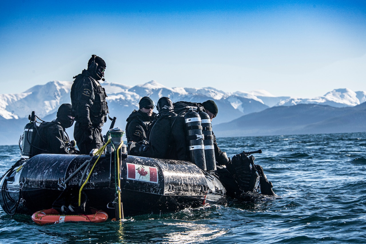 A diver in the water holds onto the side of a raft with five other personnel in it. The body of water is surrounded by snow-capped mountains.