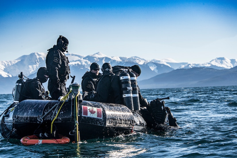 A diver in the water holds onto the side of a raft with five other personnel in it. The body of water is surrounded by snow-capped mountains.