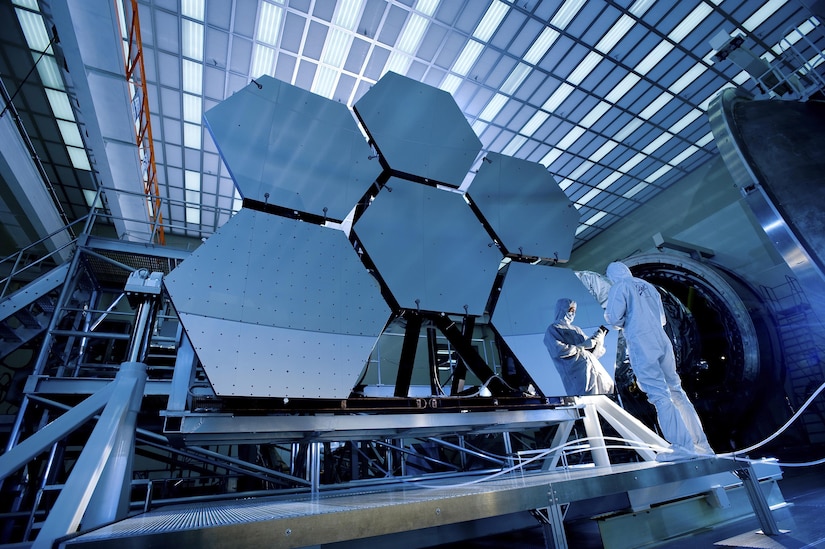 A person wearing a white protective suit stands in front of six big, hexagon-shaped mirrors.