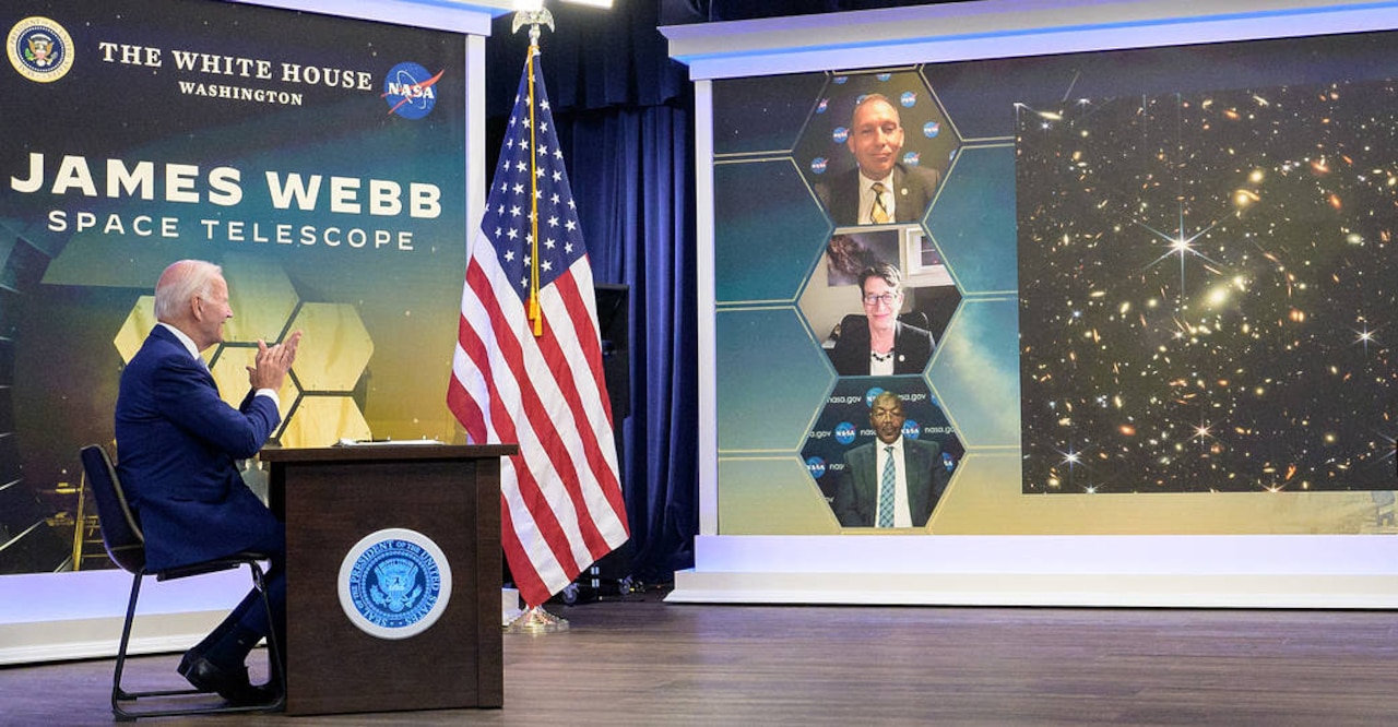 President Joe Biden teleconferences with three other people adjacent to an image of the infrared universe on a screen.