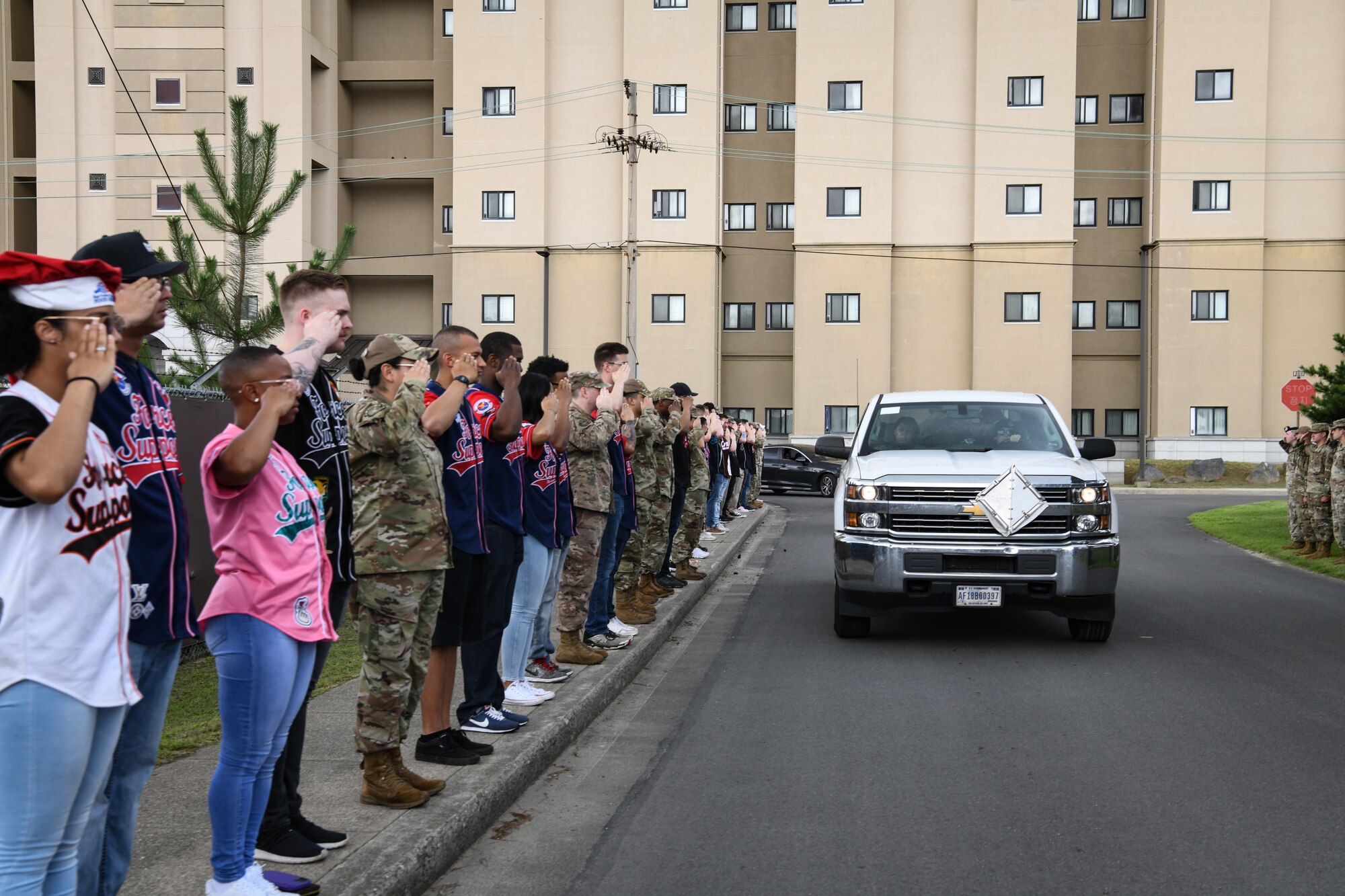 Military members in uniform and jerseys salute a pick-up truck with a military working dog in the rear.