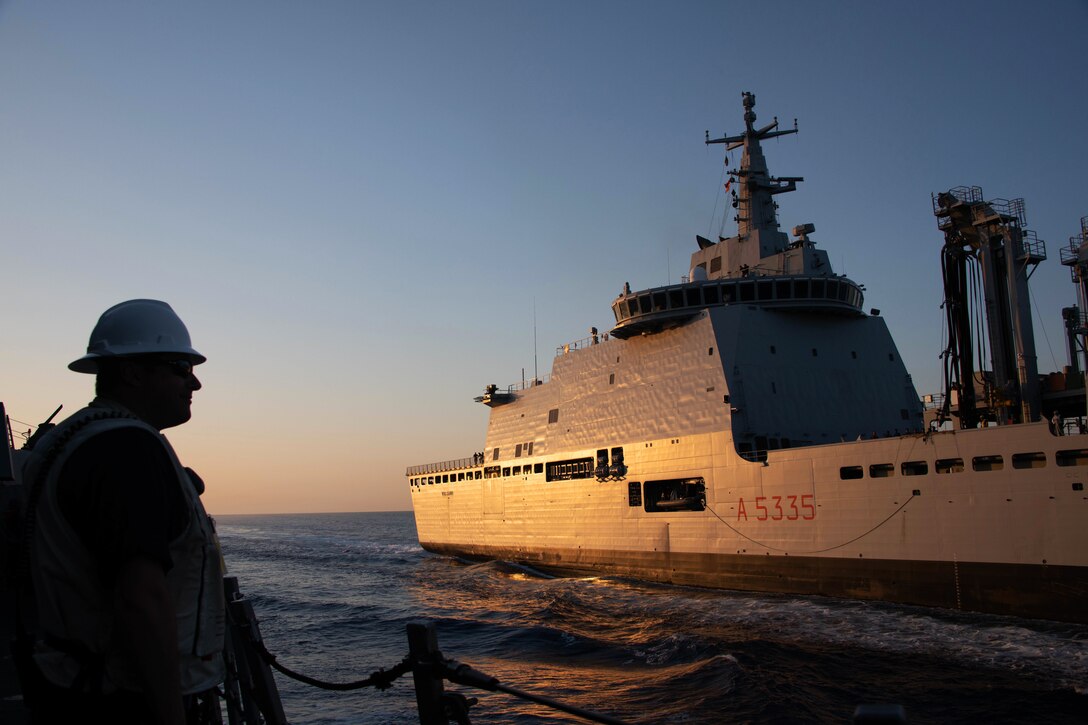 A sailor watches an adjacent ship at sail. The ship is reflecting the orange glow of the sun on the horizon.