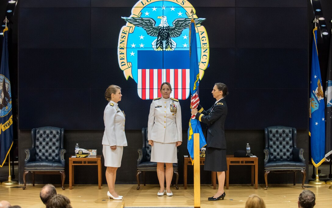 Three women stand on stage in a auditorium with flags and the DLA logo in the background. The woman on the left is dressed in Navy dress whites, the woman in the center is dressed in Navy dress whites and the woman on the right who holds the flag that was just passed from the woman on the left to the woman in the center to the woman on the right is dressed in Army dress blues.