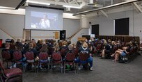 A large group of students sit in a large room watching a live-stream on a screen.