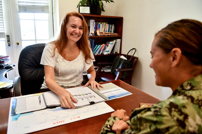 A civilian sitting at a desk talks to a service member seated on the other side.