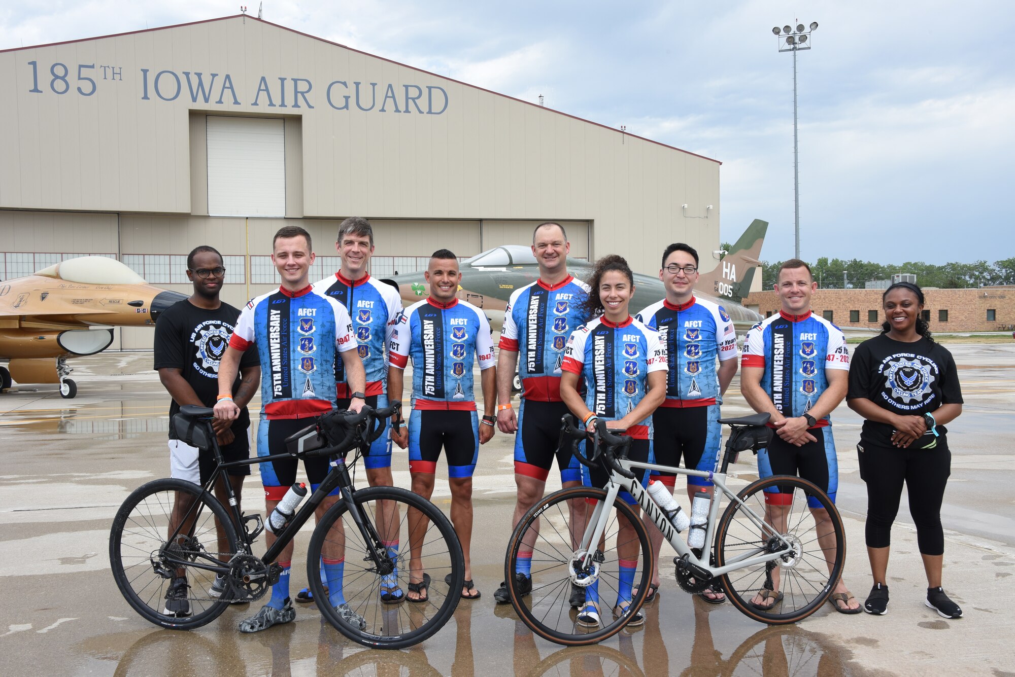 Space Force members of the Air Force Cycling Team pose for a group photo in front of a gold-painted F-16 Fighting Falcon and a F-100 Super Sabre