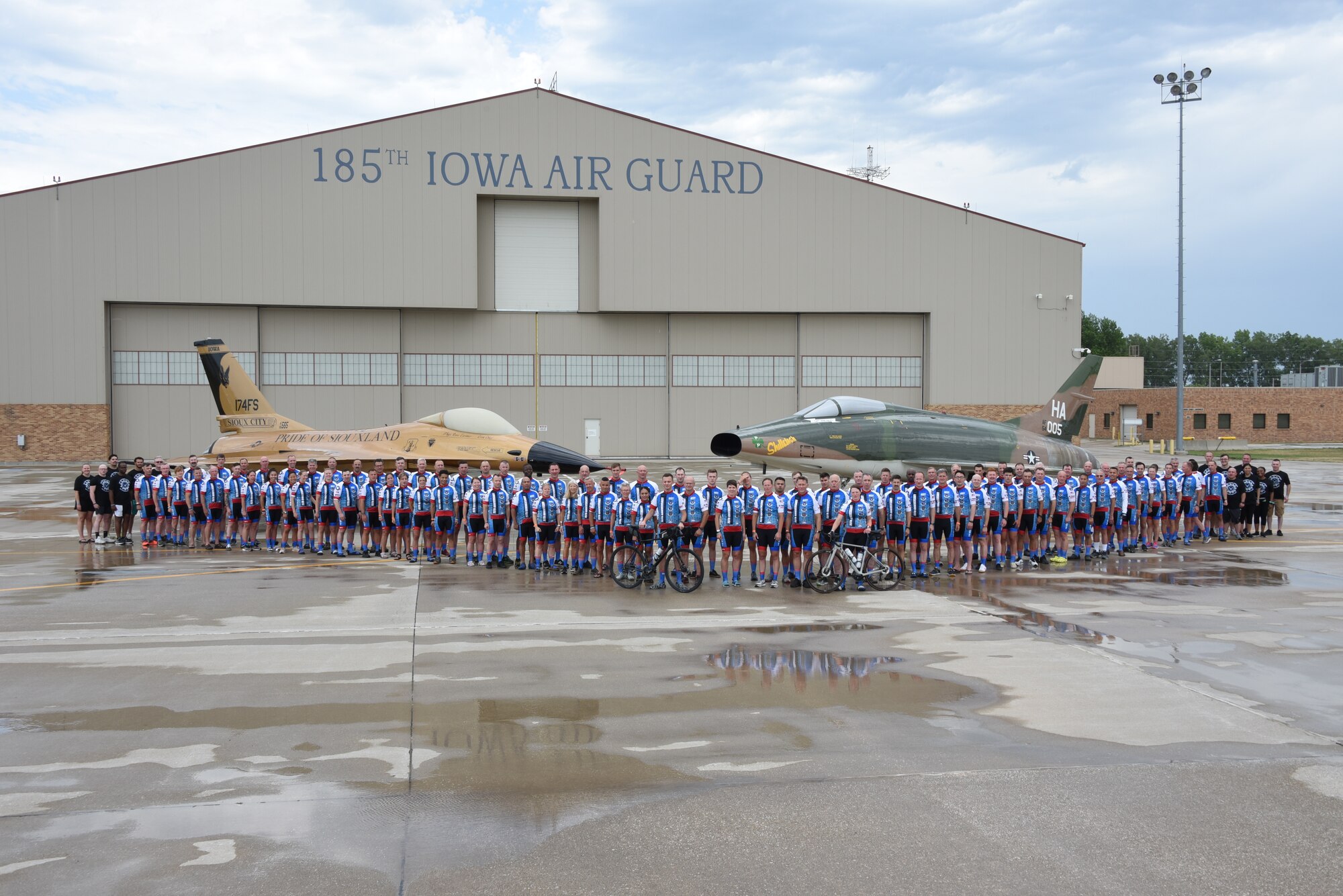 Members of the Air Force Cycling Team pose for a group photo in front of a gold-painted F-16 Fighting Falcon and a F-100 Super Sabre