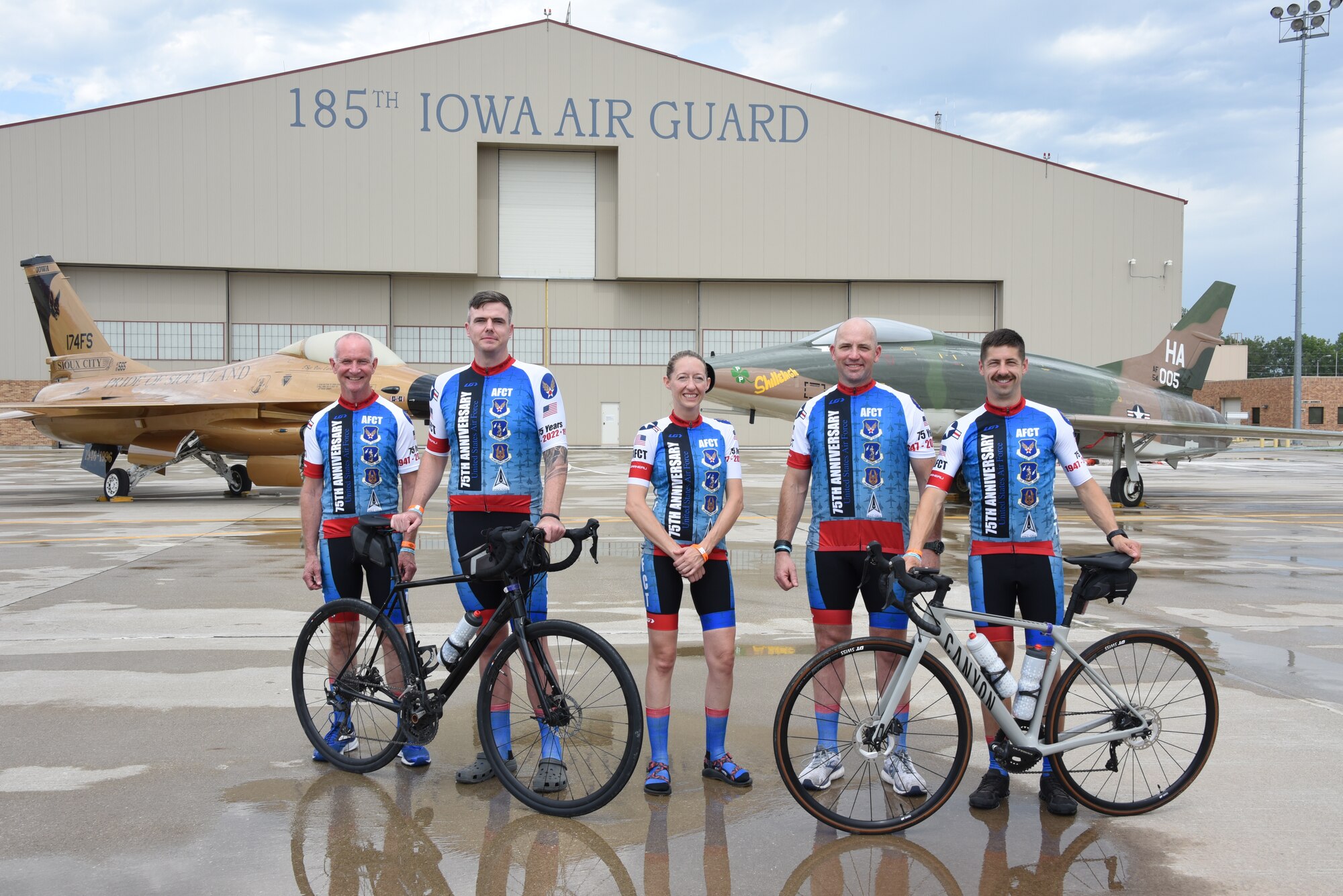 Air National Guard members of the Air Force Cycling Team pose for a group photo in front of a gold-painted F-16 Fighting Falcon and a F-100 Super Sabre
