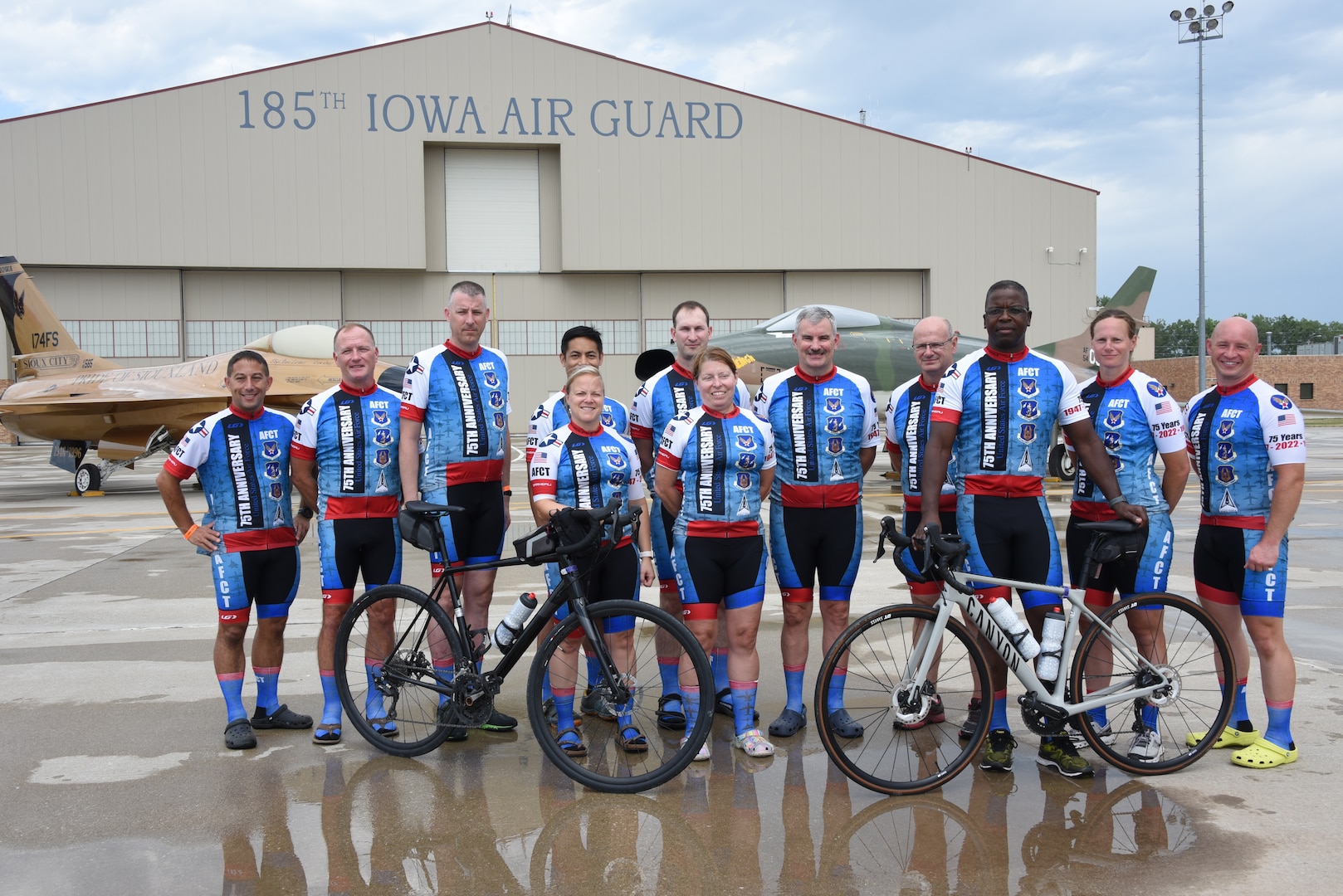 Air Force Reserve members of the Air Force Cycling Team pose for a group photo in front of a gold-painted F-16 Fighting Falcon and a F-100 Super Sabre