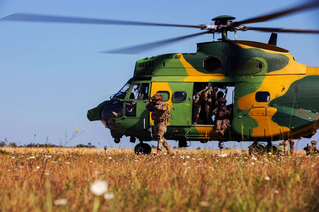 An active helicopter full of soldiers is landed in a field environment. One soldier is walking away from the helicopter’s cabin, and others can be seen on the ground behind the helicopter.
