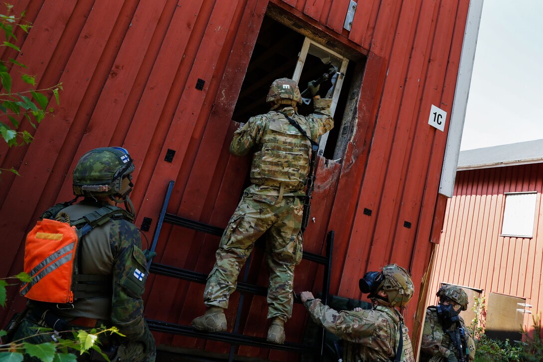 One soldier stands on a small ladder and holds a tool up into a broken window and looks inside a building while two other soldiers wearing respirator masks stand below to support the ladder. Another soldier looks from around the corner of the building.