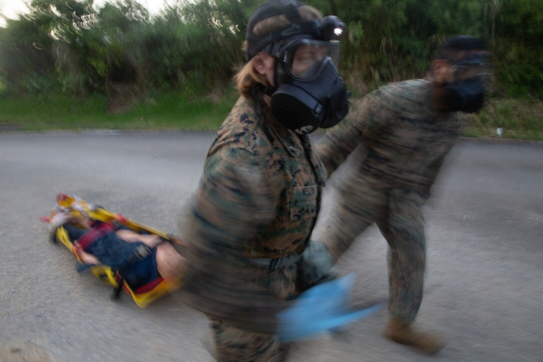 Two Marines wearing camouflage uniforms and respirators drag a bright yellow litter containing a mannequin.