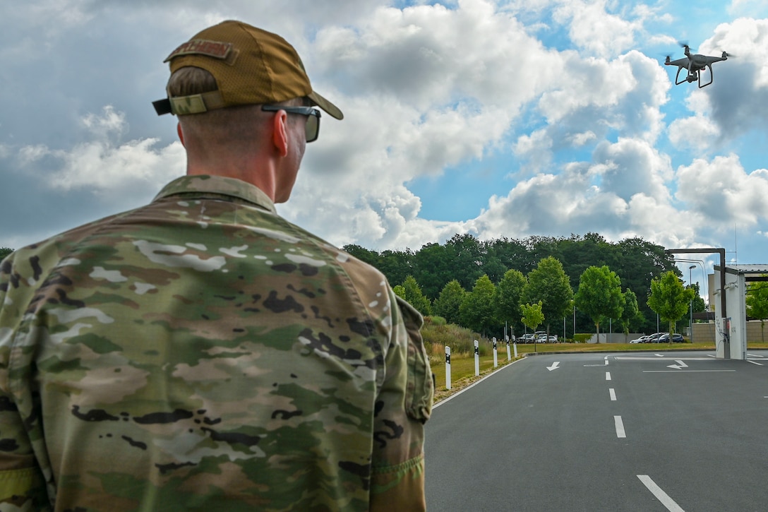 A man in camouflage uniform with his back to the camera operates a quadcopter-type drone.