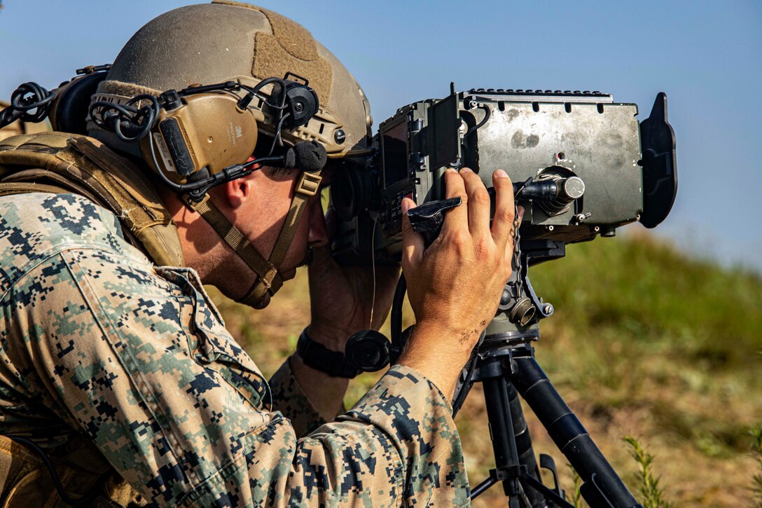 A Marine in a field looks through a device into the sky.