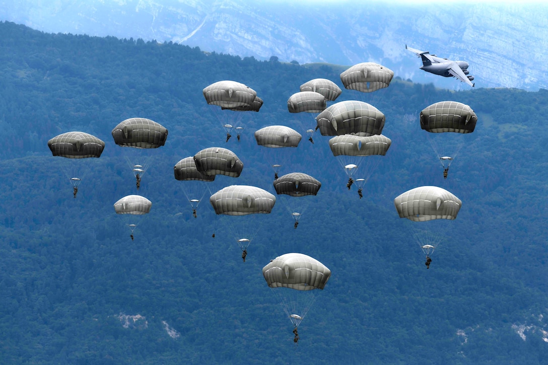 A group of soldiers descend in the sky wearing parachutes as an aircraft flies in the background.
