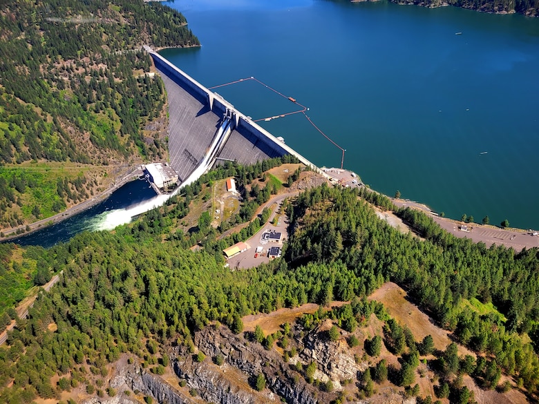 An aerial view of the Dworshak Dam, in Orofino, Idaho. It is a concrete gravity dam. The water held in the reservoir is a deep cerulean blue. A large plume of white water pours over from the spillway.