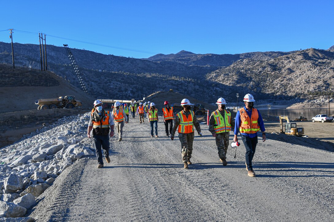 A group of people in hardhats and safety vests walk on the graveled top of an earthen dam. Behind them is an active construction site and mountains in the distance.