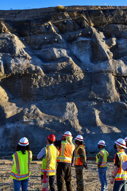 a group of people in hardhats and safety vests looks up at a wall of rock that has been carved out of a mountain by blasting.