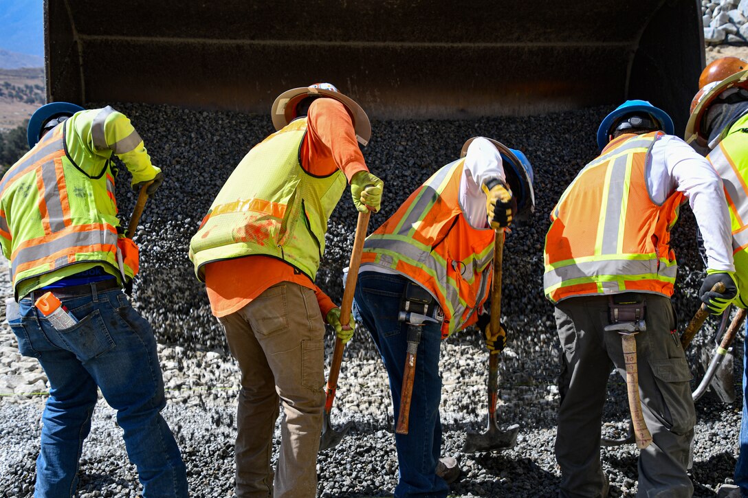 five men stand in a line, shoveling gravel while a front end loader dumps it on the dam in front of them.