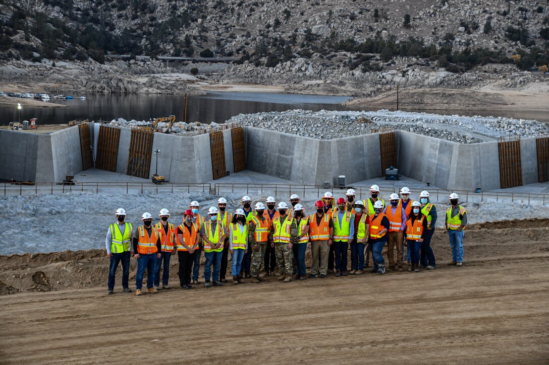 A group of people stand in front of a construction site. Behind them is an expanse of blasted rock and a labyrinth weir. Further back is a lake and mountains.