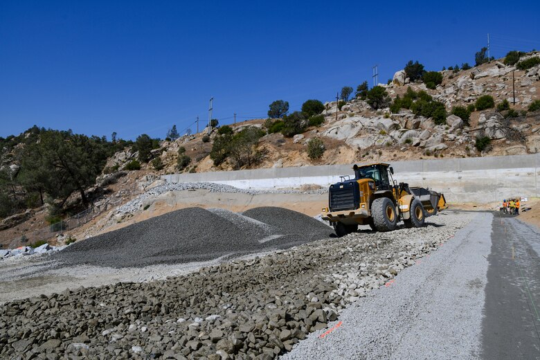 Looking down an earthen dam under construction with a road and mountains in the distance, a front end loader carries gravel to place on the dam.