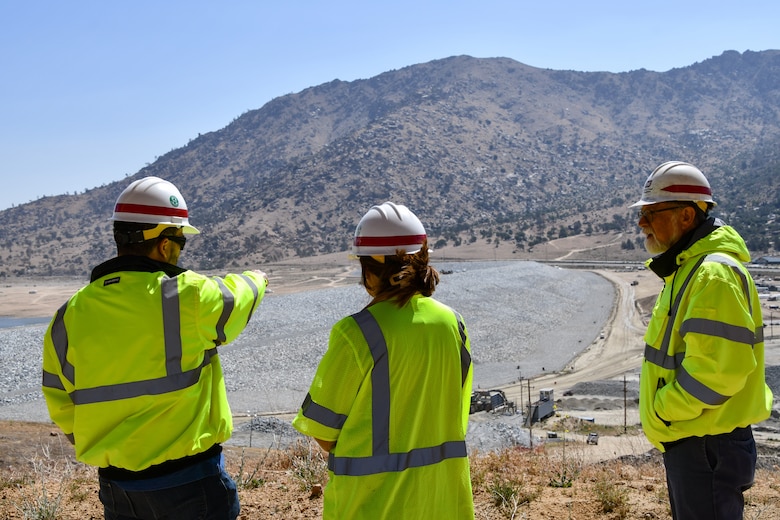 Two men and one woman overlook an earthen dam with mountains in the distance. The woman is in the middle with the two men on either side. The man on the left is pointing out to the dam, and the others are listening to him speak.