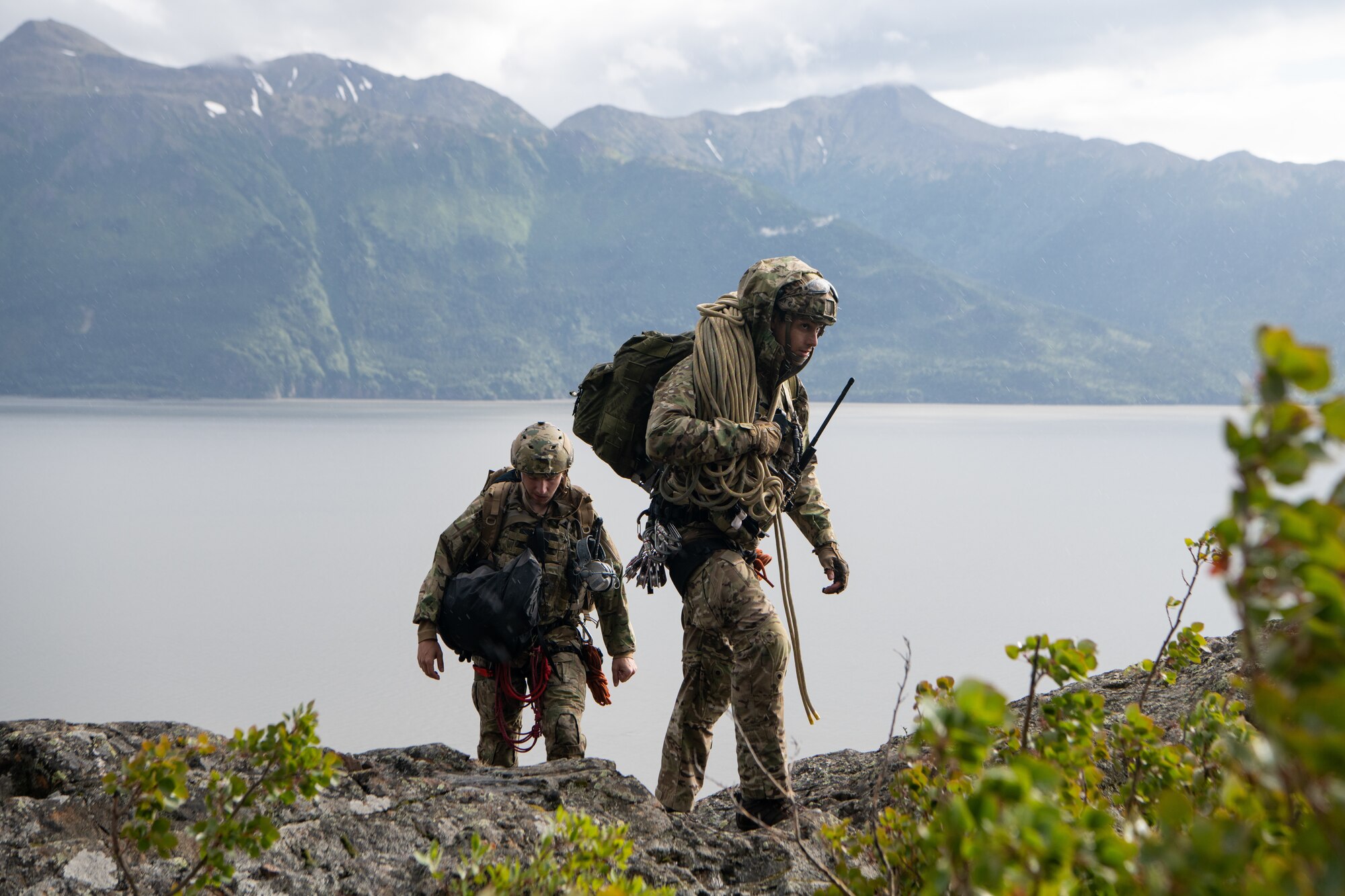 Two Airmen carry climbing gear over rocky terrain.