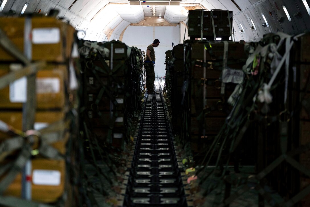 A man stands in cramped quarters and looks at a cases stacked in rows in front and behind him.