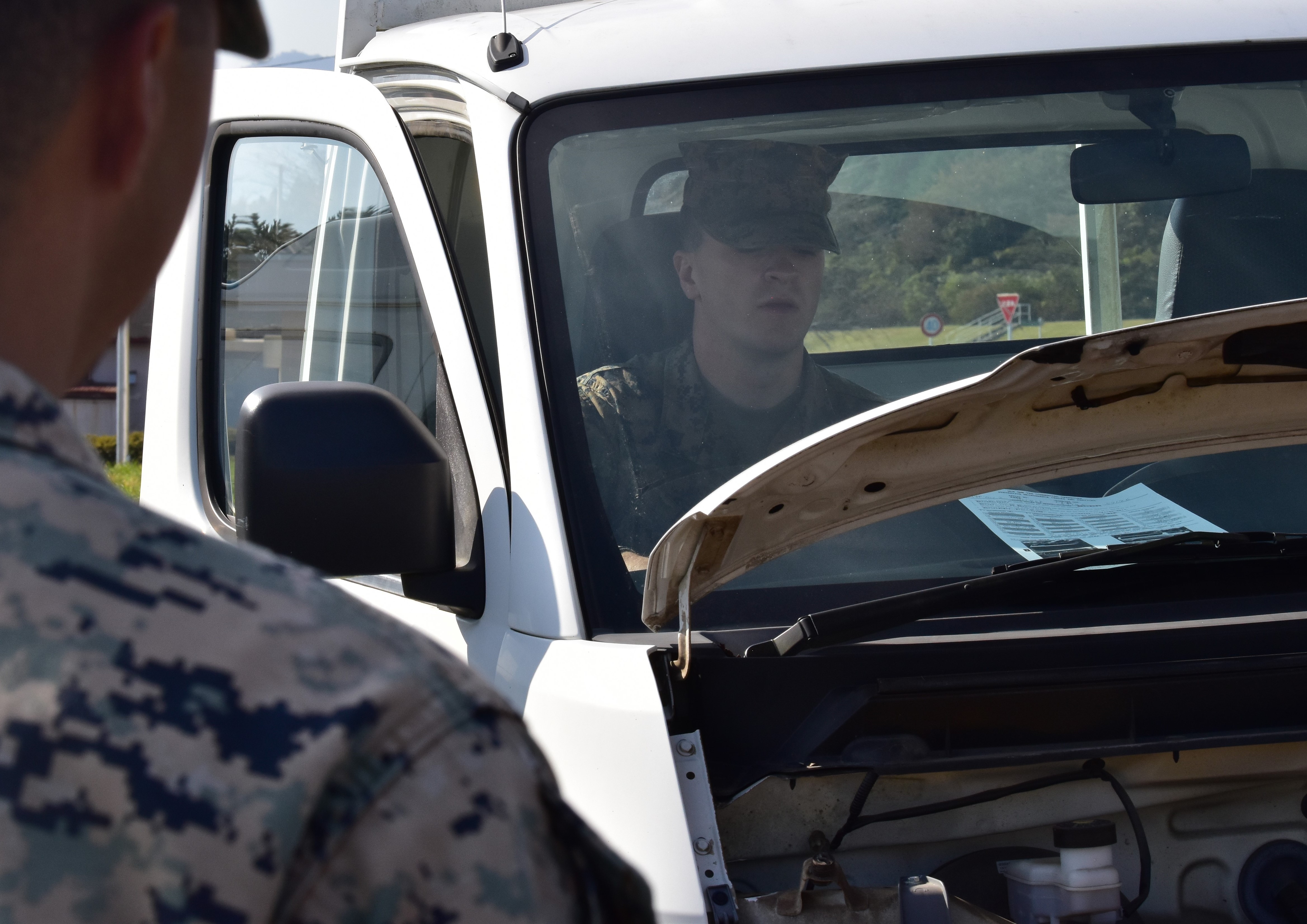 A Marine's head from behind looking at another Marine in the passenger seat of a white vehicle with the hood up.