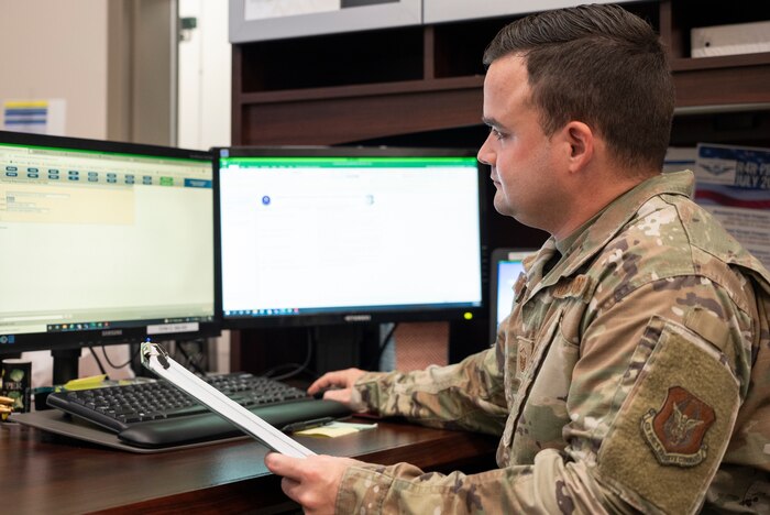 Airman with clipboard in hand looks at computer.