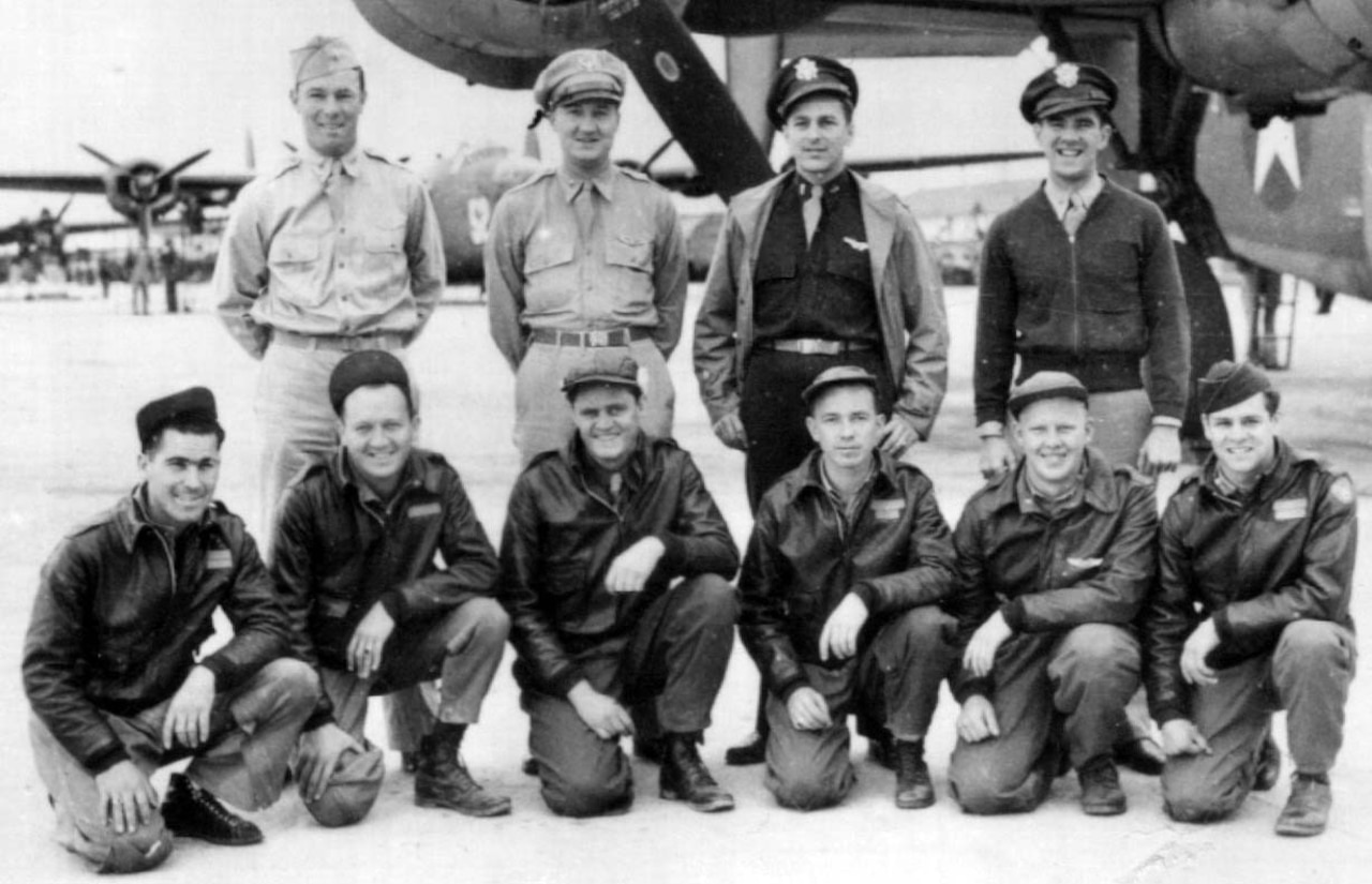 Ten men pose in two rows for a photo beside an airplane.