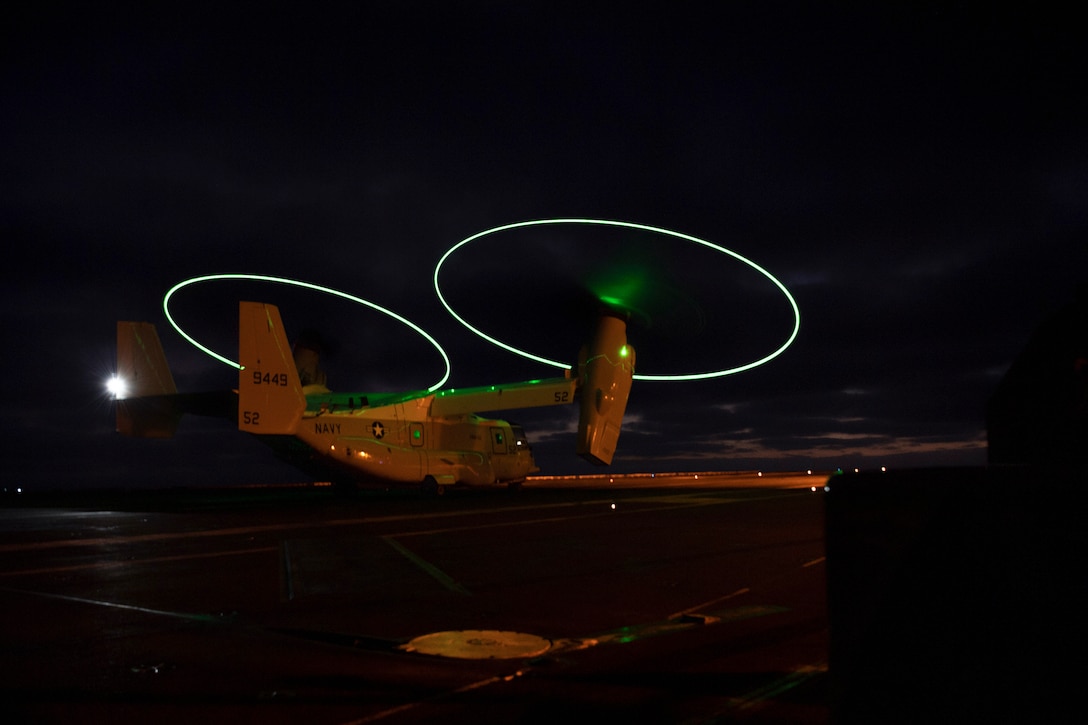 A Navy aircraft takes off in the dark from a ship at sea illuminated by green lights.